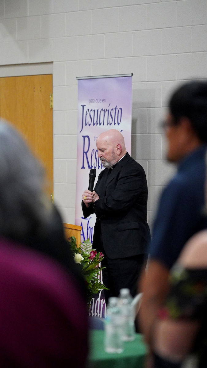 Bald man in black suit prays with a microphone, near banner with "Jesucristo". Floral arrangement and water bottles visible. Indoor setting.