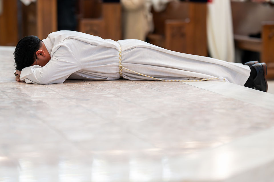 Person in white robe lies prostrate on polished marble floor in a church setting, conveying a solemn and reverent mood.