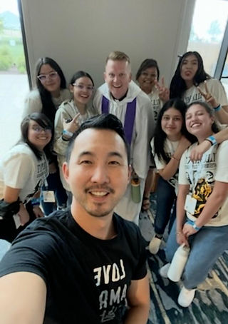 Group of smiling people taking a selfie indoors. One person wears a purple stole. Others in matching shirts. Casual and cheerful mood.