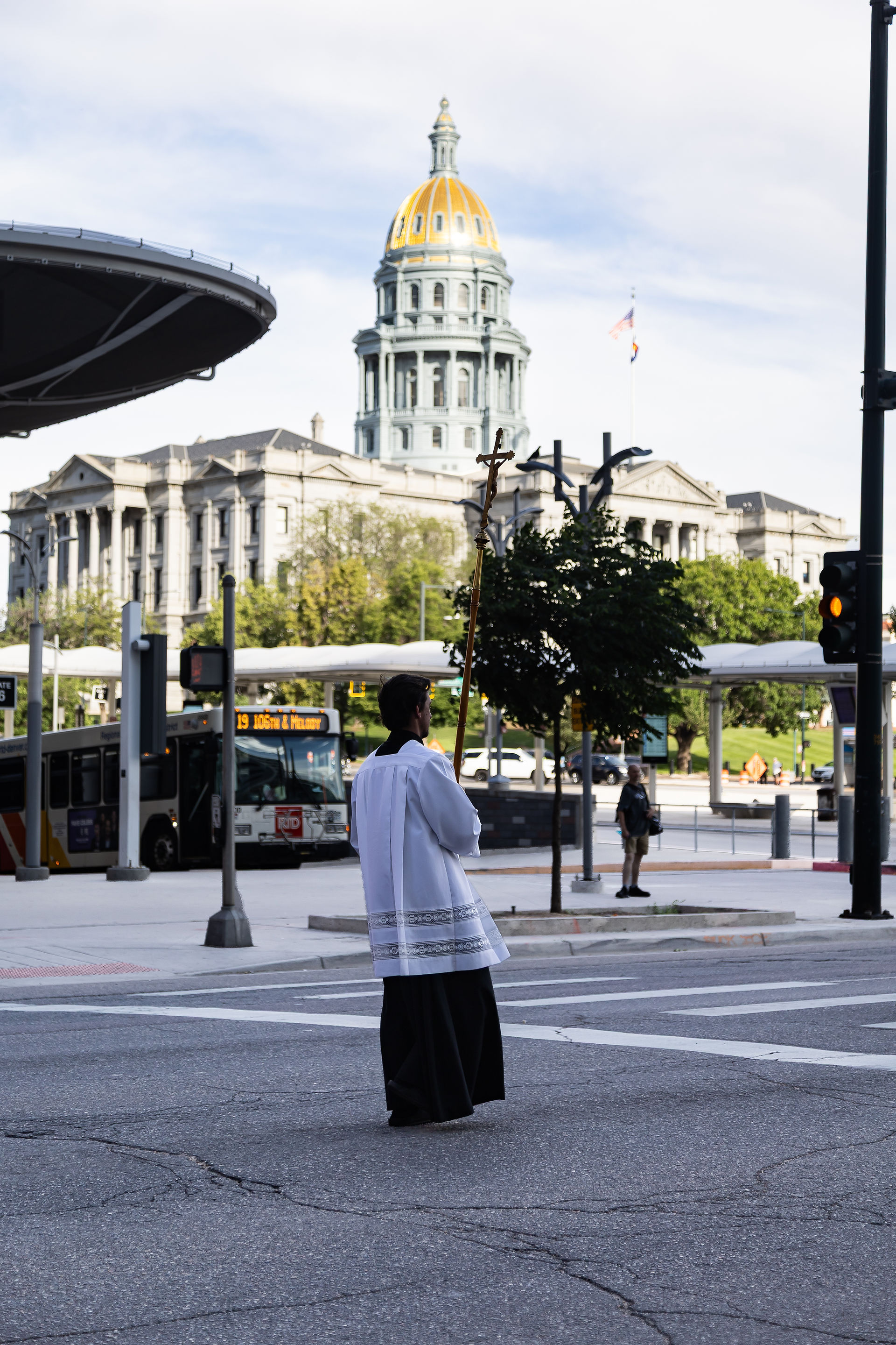 Person in white religious vestments holds a cross on a city street, with a gold-domed capitol building and bus behind him.