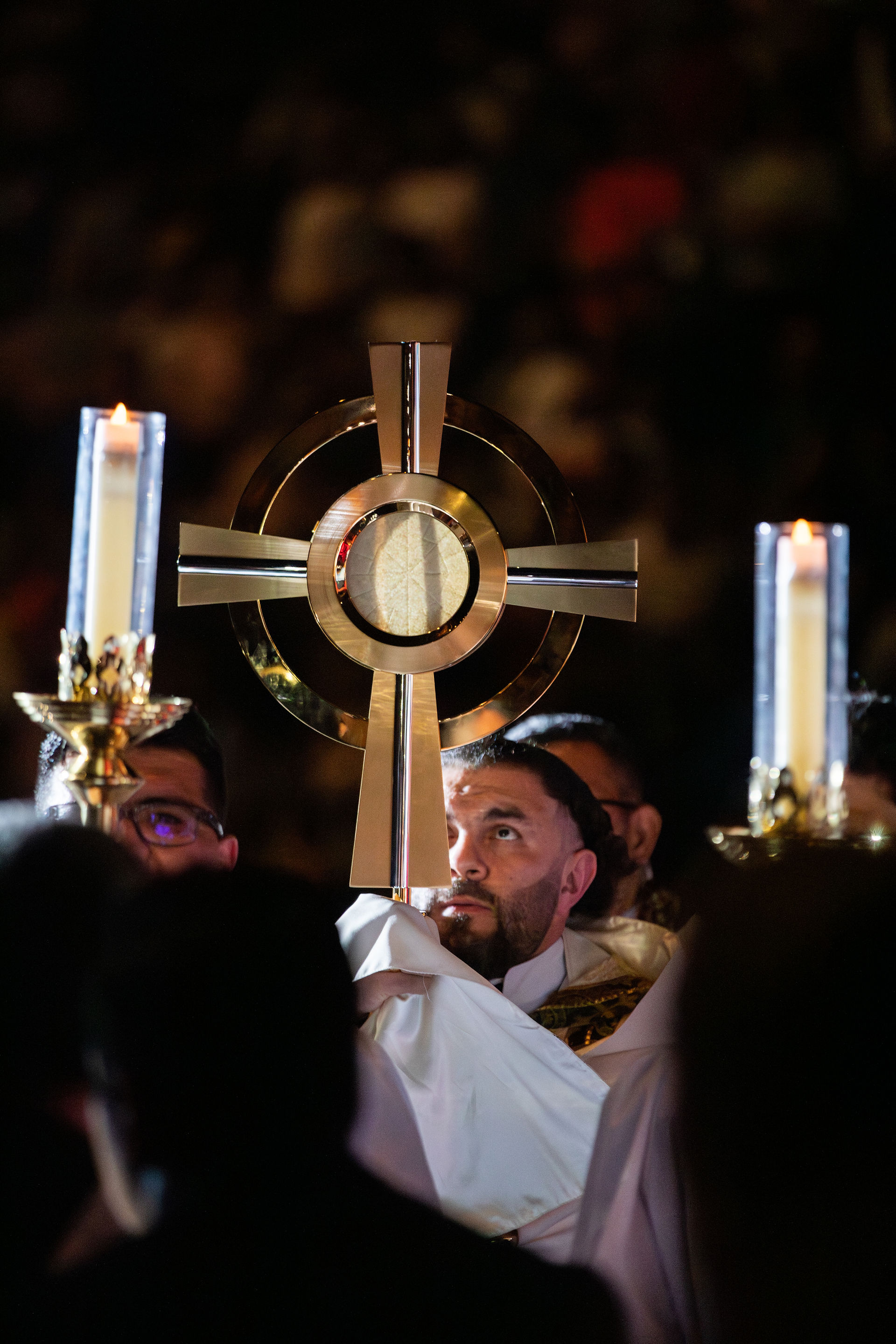 A gold monstrance with a visible Eucharistic host stands between two lit candles in a dimly lit church setting, highlighting a solemn religious ceremony.