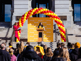 Speaker at podium under red and yellow balloon arch, addressing crowd on steps. Background features gray stone building. Text: "School Choice."