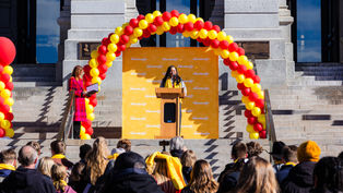 Speaker at podium under red and yellow balloon arch, addressing crowd on steps. Background features gray stone building. Text: "School Choice."