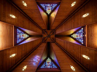 Wooden ceiling of a church with triangular stained glass panels casting colorful light. Hanging lamps and a fan are also visible.