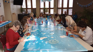 People sitting around a long table with a sky-themed blue cover, writing on sheets. "Welcome" banner and plants decorate the room.