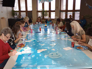 People sitting around a long table with a sky-themed blue cover, writing on sheets. "Welcome" banner and plants decorate the room.