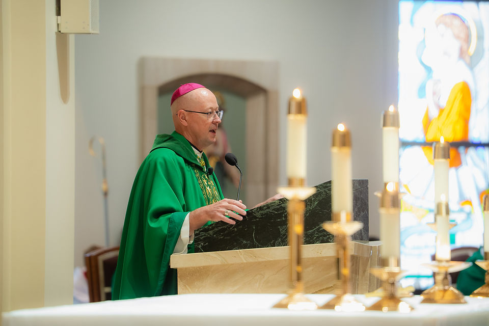 Bishop in green vestments speaks at a church lectern beside lit candles, with stained glass glowing in the background.