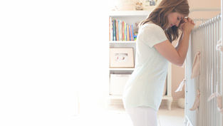 A woman leans on a crib in a softly lit nursery, eyes closed, appearing thoughtful. Books and framed photos fill shelves in the background.