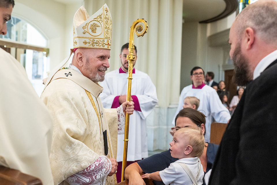 A bishop in ornate robes with a gold staff smiles at a child held by a woman in a church. Clergy and congregation are visible in the background.