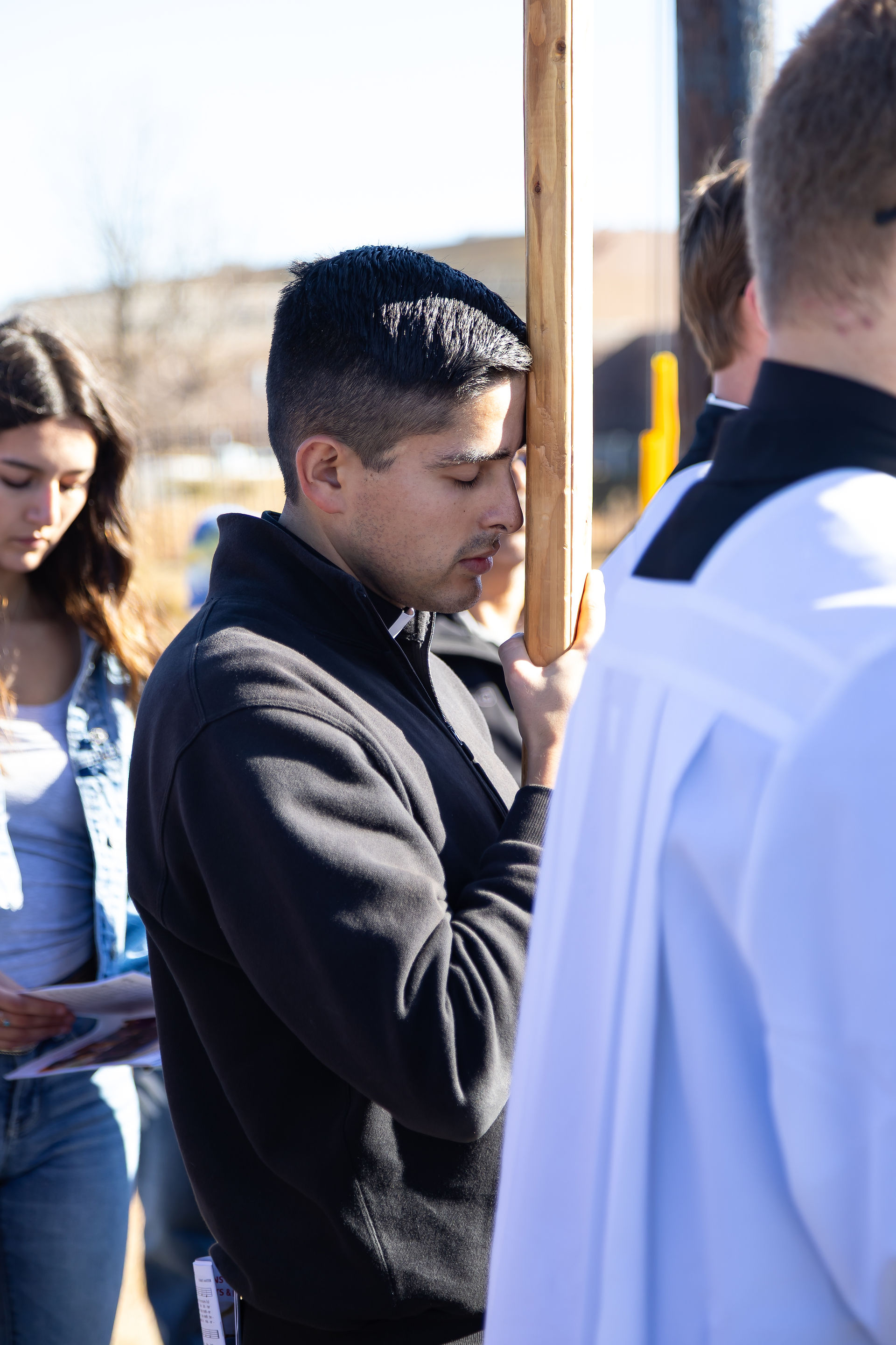 Man in black jacket prays with a wooden cross, closed eyes. Others nearby, one holding paper. Outdoor, sunny setting, serious mood.