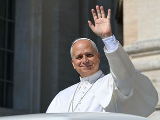 Pope in white robes smiles and waves from a balcony against a stone building backdrop, conveying a warm, welcoming mood