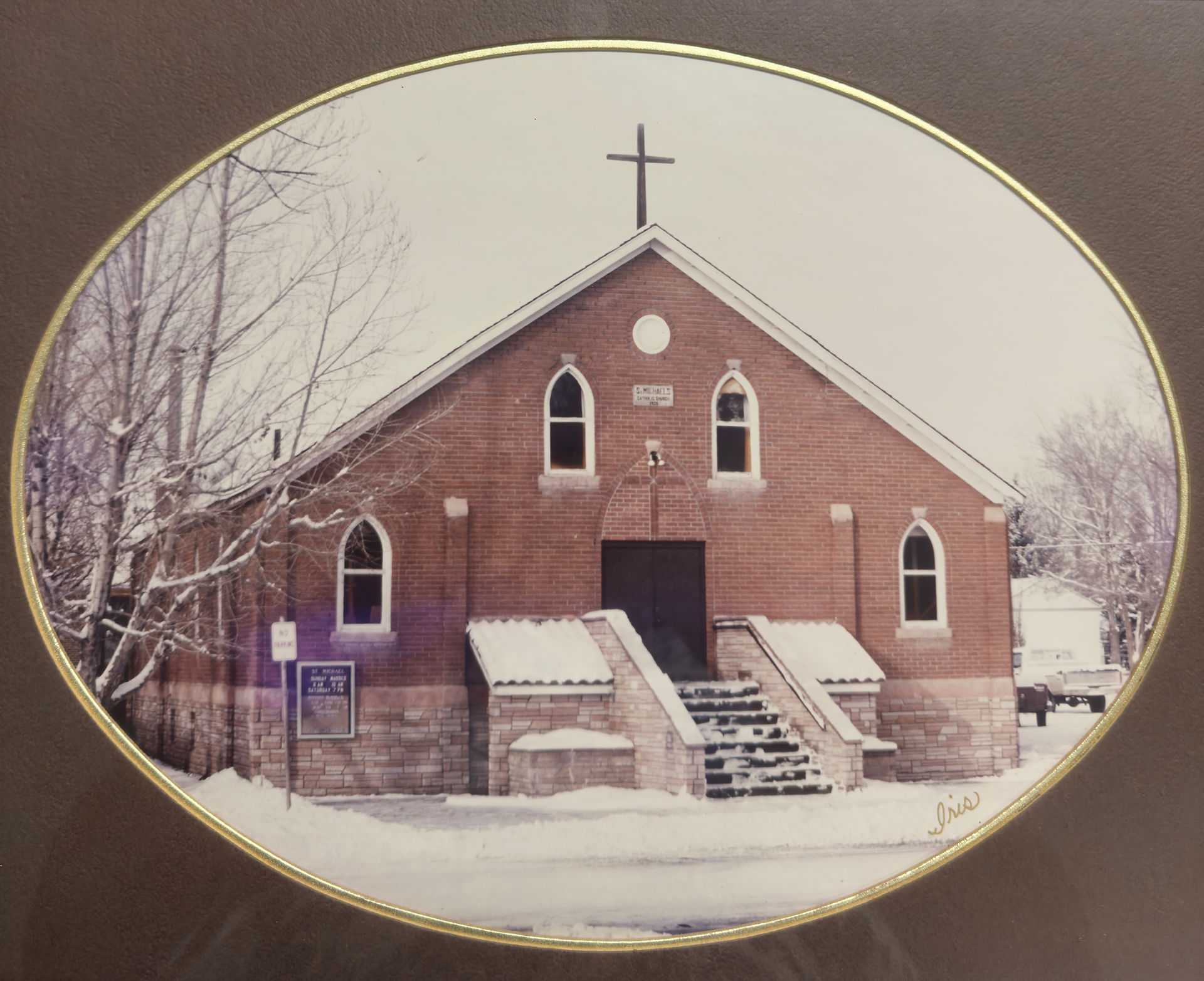 Red brick church with snow-covered steps, surrounded by bare trees. A sign reads "St. Michael's." A peaceful winter scene.