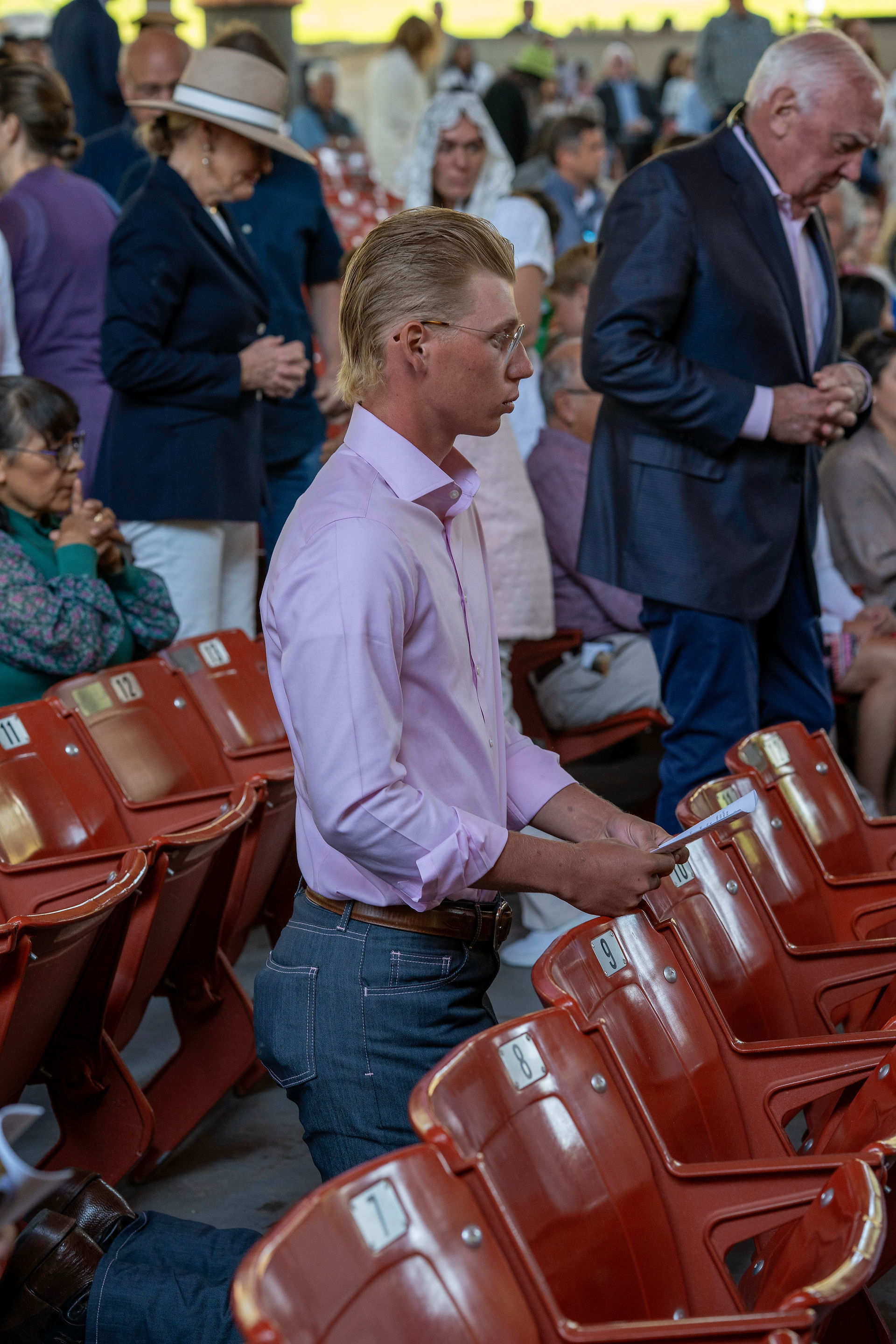 A person in a light purple shirt and jeans kneels between rows of red seats in an indoor gathering, surrounded by others who are seated or standing.