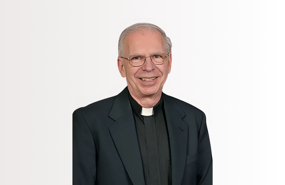 Smiling man in clerical attire with a white collar and glasses, set against a plain white background.