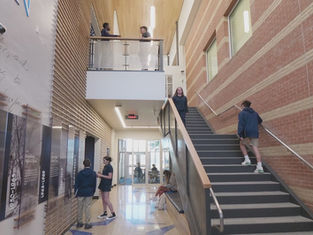 Students in a school hallway, some standing, some walking. Brick walls, staircase, and informational panels create a modern academic setting.
