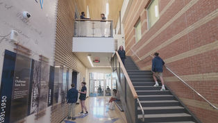 Students in a school hallway, some standing, some walking. Brick walls, staircase, and informational panels create a modern academic setting.
