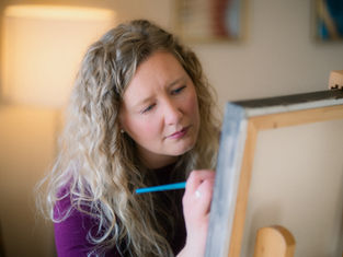 Woman with curly hair painting on a canvas in a warmly lit room, focused expression, blue brush in hand, wearing a purple top.