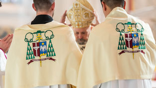Two clergy in decorated ivory vestments stand with a bishop holding a mitre. Symbols and Latin text are visible. Reverent setting.