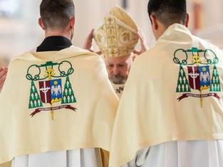 Two clergy in decorated ivory vestments stand with a bishop holding a mitre. Symbols and Latin text are visible. Reverent setting.