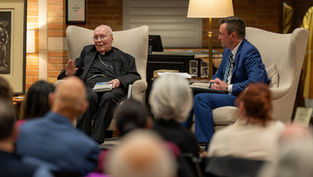 Two men sit in armchairs having a conversation. One holds a book in a warmly lit room. An audience listens attentively in the foreground.