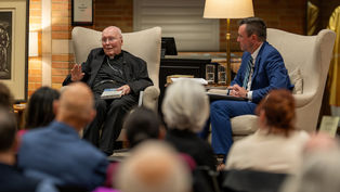 Two men sit in armchairs having a conversation. One holds a book in a warmly lit room. An audience listens attentively in the foreground.