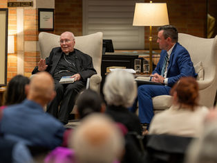 Two men sit in armchairs having a conversation. One holds a book in a warmly lit room. An audience listens attentively in the foreground.