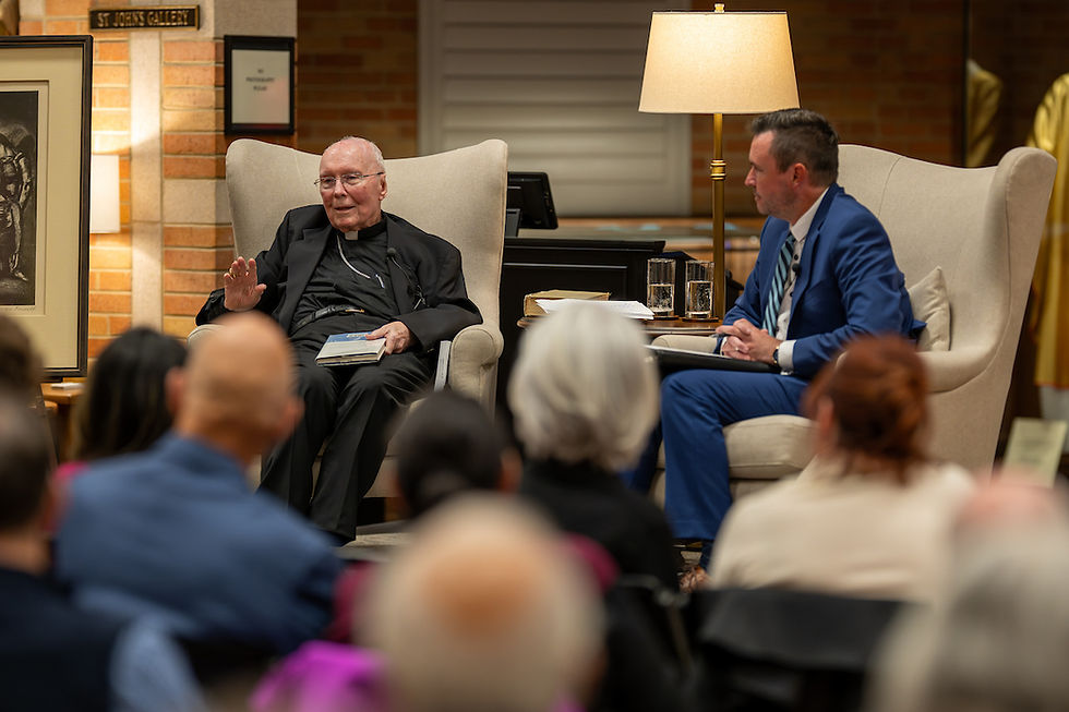 Two men sit in armchairs having a conversation. One holds a book in a warmly lit room. An audience listens attentively in the foreground.