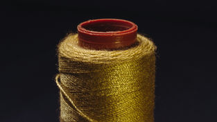 Close-up of a spool of golden thread with a deep red top against a dark background, highlighting the thread's sheen and texture.