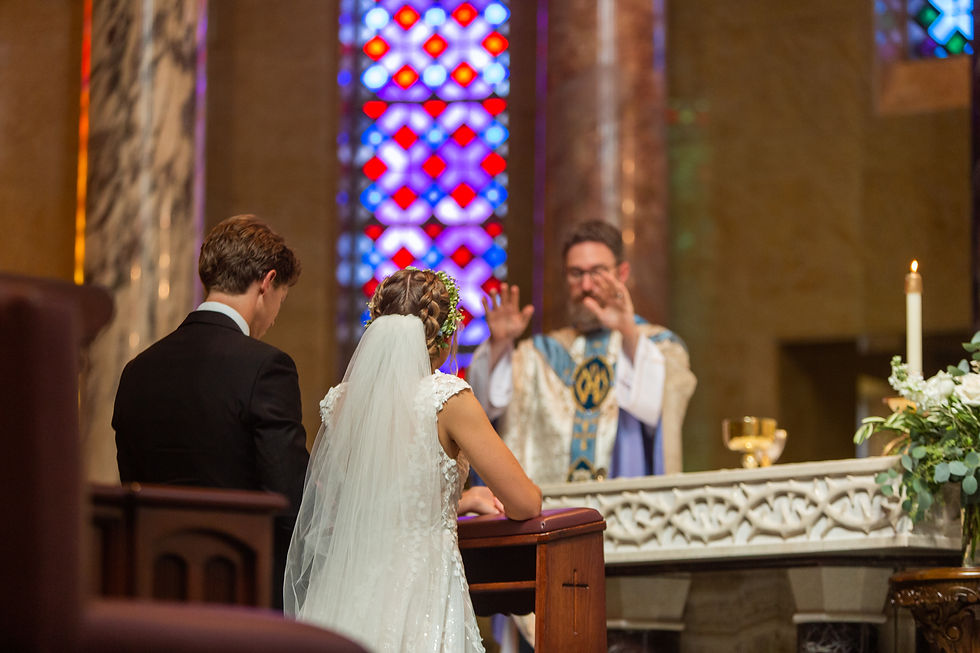 Bride and groom kneel before priest in colorful church with stained glass. Priest raises hands in blessing. Candle and flowers present.