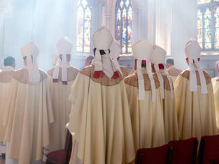 Clergy in cream robes and mitres stand in a church with stained glass. Sunlight filters in, creating a serene atmosphere.