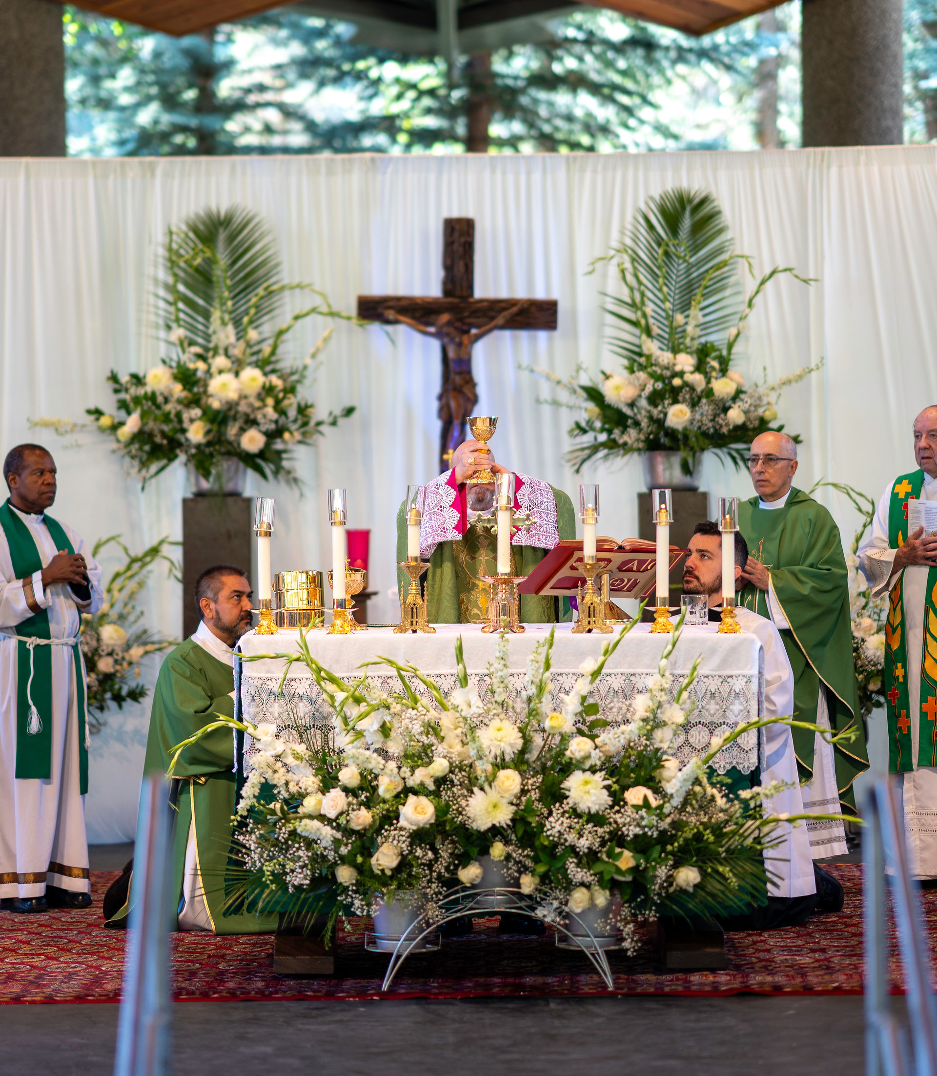 Priests celebrate Mass, holding a chalice at an altar adorned with candles and white flowers. A crucifix and greenery are in the background.