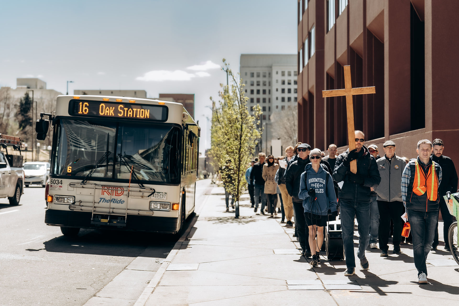 A group processes along a city sidewalk beside a bus, led by a man carrying a wooden cross.