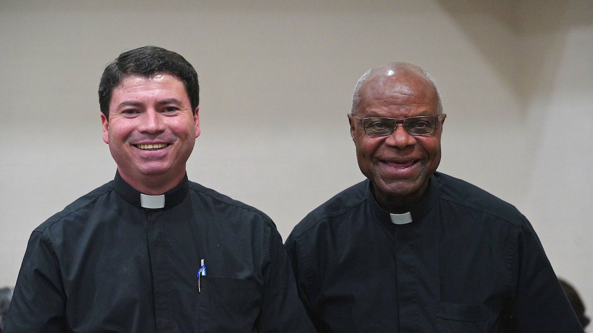 Two smiling clergymen in black clerical shirts stand in a neutral indoor setting. One has glasses, and another holds a pen in his pocket.
