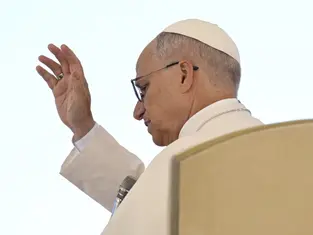 A person in white attire raises their hand in a blessing gesture. The background is plain, creating a serene and solemn atmosphere.