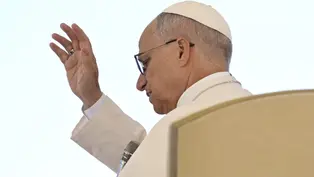 A person in white attire raises their hand in a blessing gesture. The background is plain, creating a serene and solemn atmosphere.
