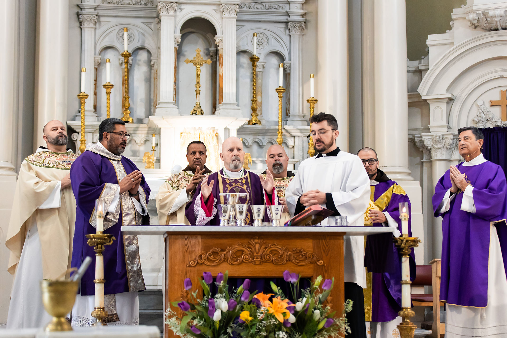 Clergy in purple and white vestments stand at an altar, praying in a ornate church. Candles and flowers decorate the scene.