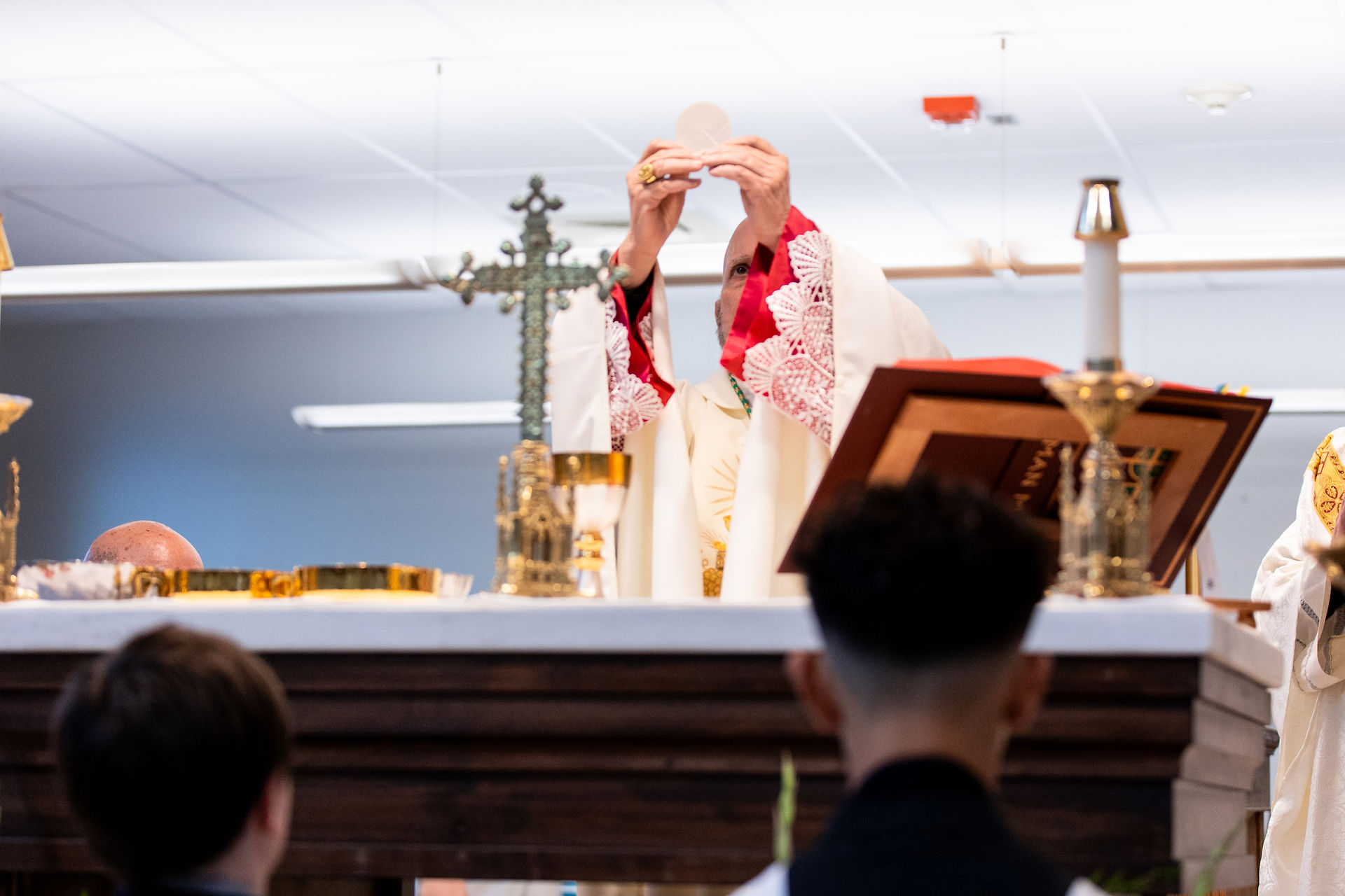 Bishop in white and red vestments holds up a host at altar during service. Ornate cross and chalice visible, with people in attendance.