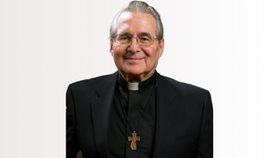 Elderly man in a black clerical suit with a white collar and cross necklace, smiling against a plain background.