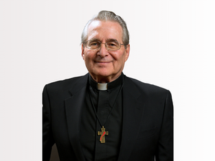 Elderly man in a black clerical suit with a white collar and cross necklace, smiling against a plain background.