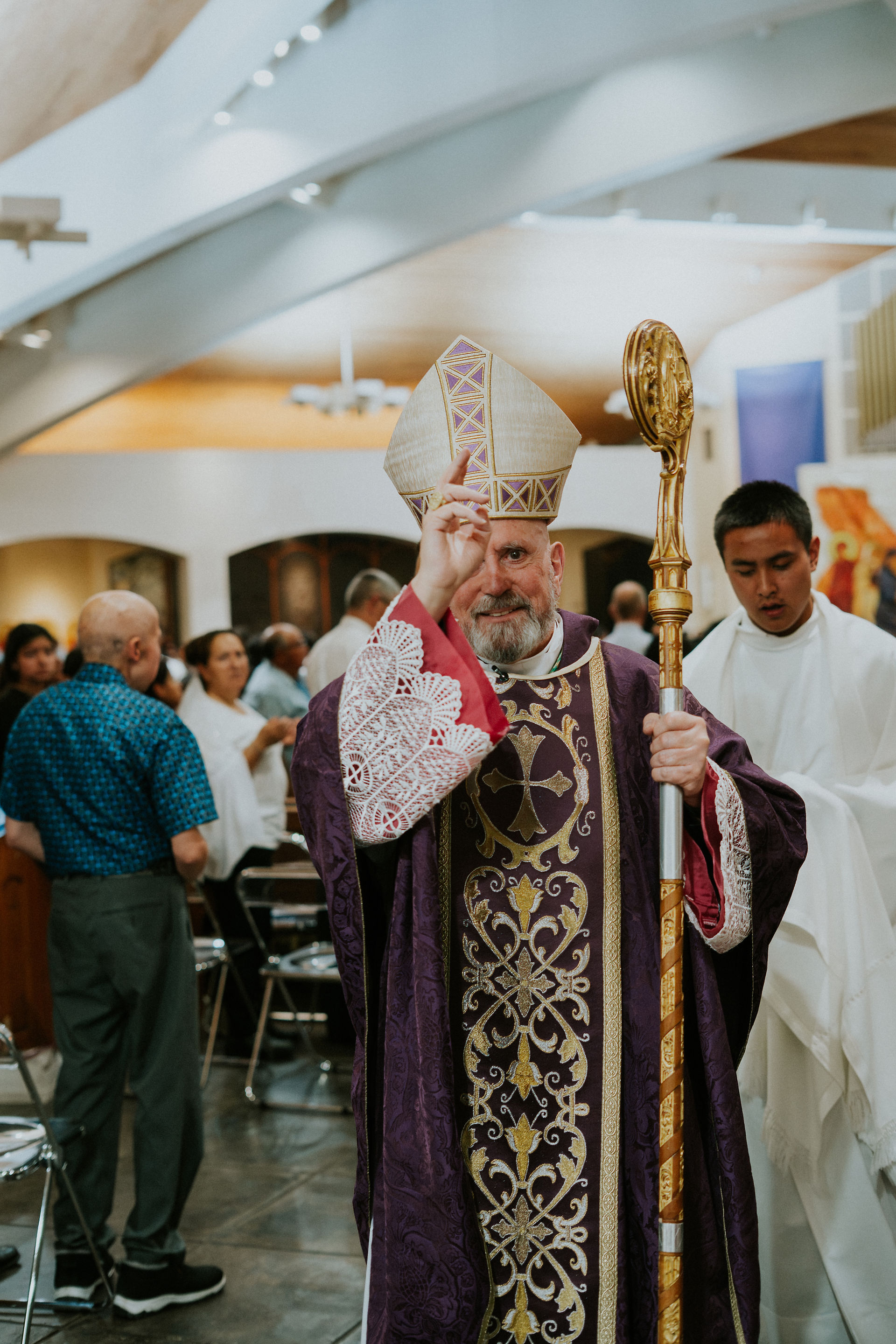 A bishop in ornate purple robes and mitre raises a hand in blessing, holding a staff. Background: church interior with people gathered.