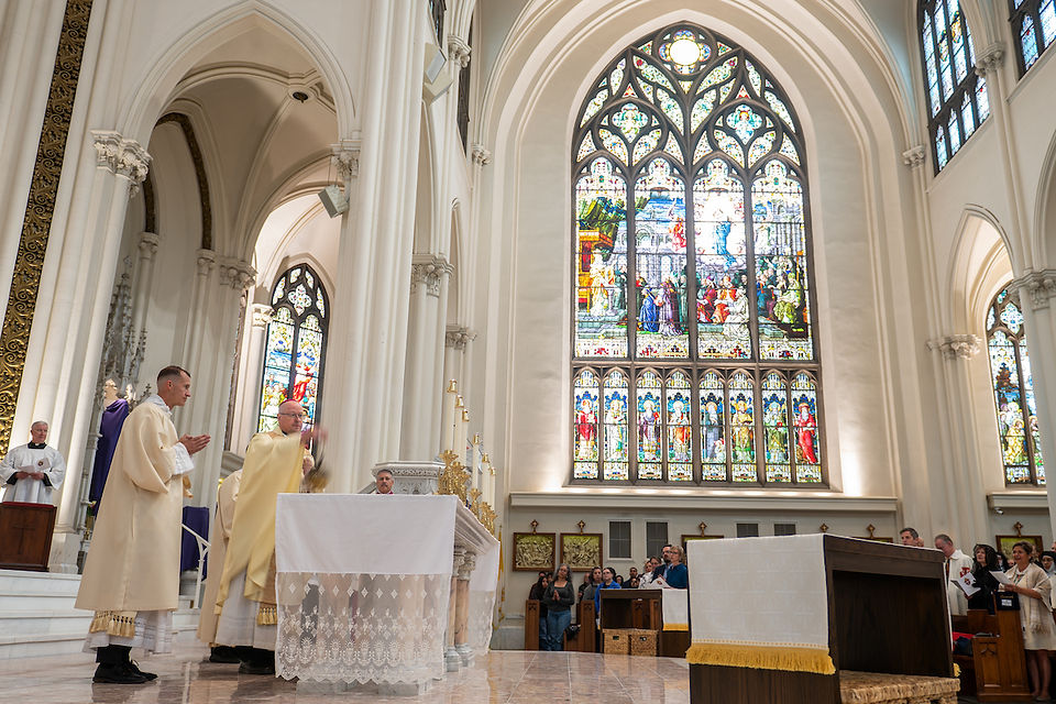 Clergy in cream robes conduct a ceremony in a church with tall arches and a colorful stained glass window. Congregation seated, solemn mood.