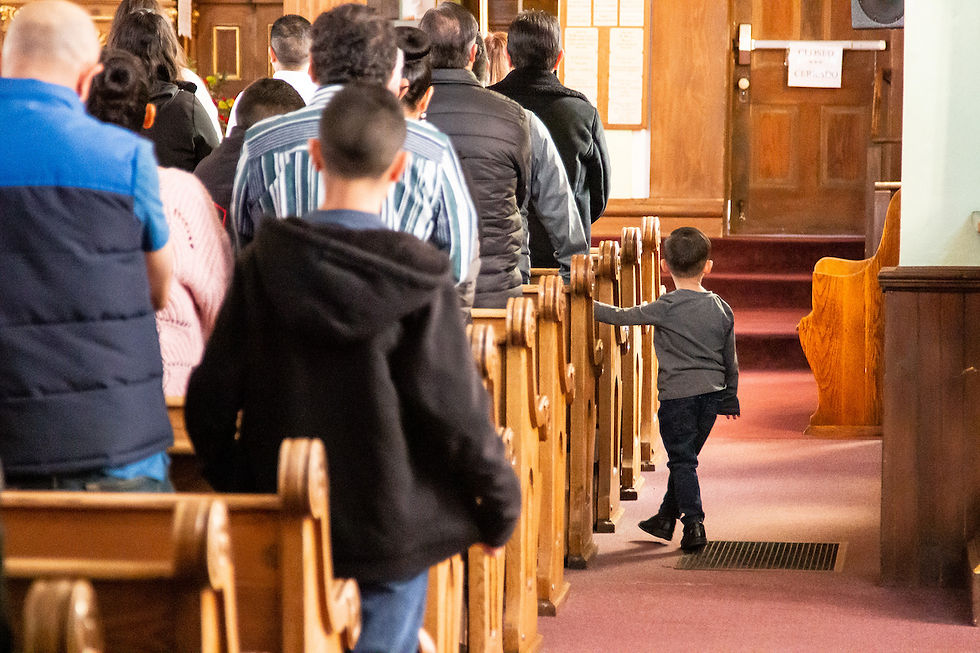 People stand in a church aisle facing the altar. A child walks beside wooden pews. Warm lighting and a wooden door are visible.