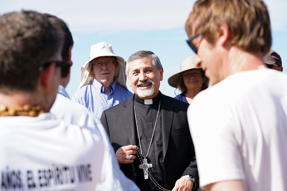 Smiling priest in black clerical clothes with a cross necklace talks with a small group outdoors under a bright blue sky.