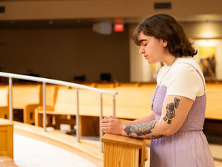 Young person in a lavender dress and tattoos prays with eyes closed in a quiet, wooden pew-filled room, exuding tranquility.