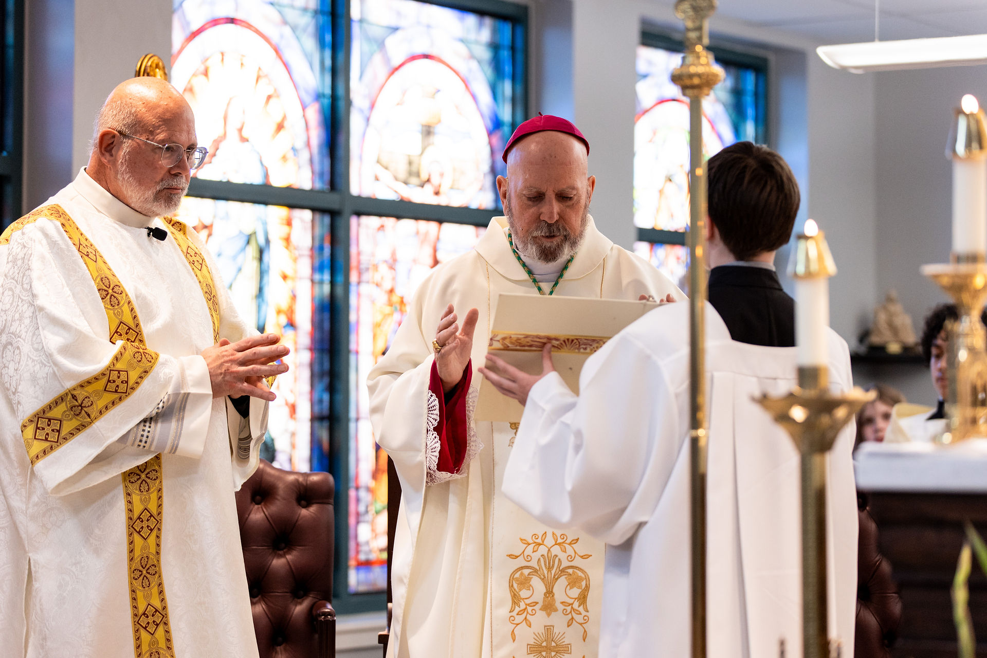 Three clergy in ceremonial robes stand in a church with stained glass windows, engaged in a religious service. Candles add ambient light.