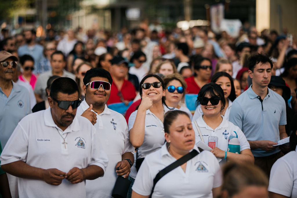 Crowded outdoor gathering of people in white polos and sunglasses, some smiling and waving in a sunny street.