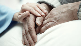 Hands of an elderly person being gently held by a younger person on a white blanket, conveying warmth and care in a soft-focus setting.