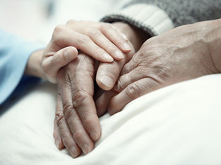 Hands of an elderly person being gently held by a younger person on a white blanket, conveying warmth and care in a soft-focus setting.