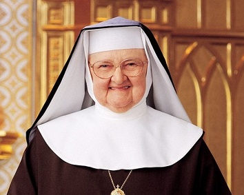 Smiling nun in black-and-white habit and glasses stands indoors against a warm wooden background.