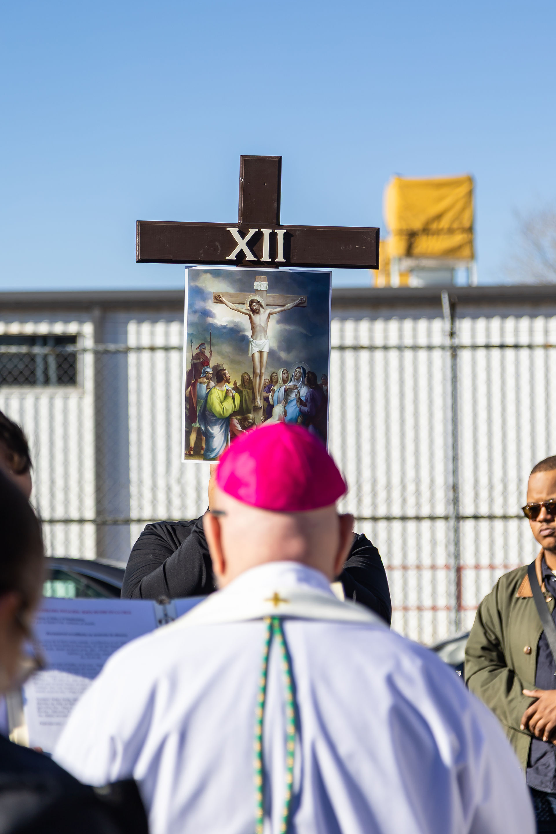 Clergyman with pink hat facing a group. A cross labeled "XII" with a crucifixion image is held. Outdoor setting with a clear sky.